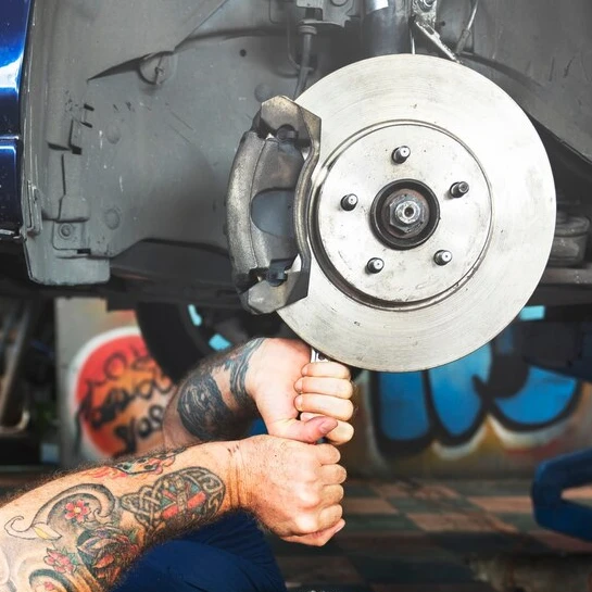 A tattooed mechanic in Auto Repair Surrey and Delta skillfully works on a car's brake system, hands gripping a tool to tighten or loosen components.