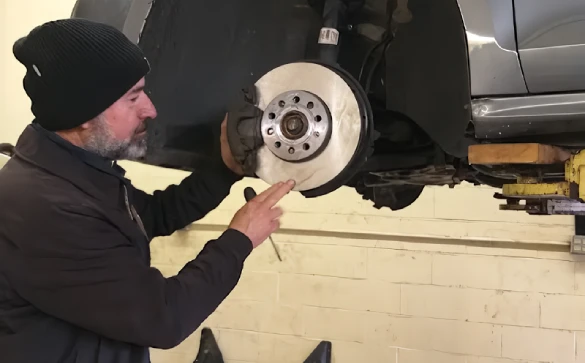 A mechanic wearing a black beanie and jacket examines the brake disc of a car lifted in an Auto Repair Surrey and Delta garage.