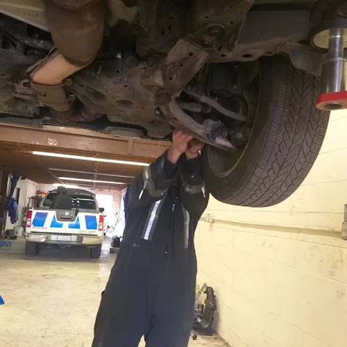 A mechanic wearing a black jumpsuit works under the raised front end of a vehicle in a garage. A white pickup truck is visible in the background, emblematic of the reliable service at Auto Repair Surrey and Delta.