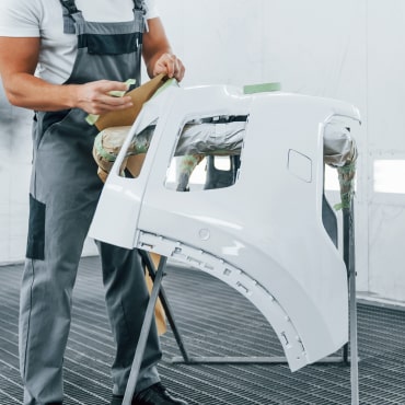 A person wearing gray overalls sands the surface of a white car bumper in an auto repair shop in Surrey and Delta. The bumper is positioned on a stand.
