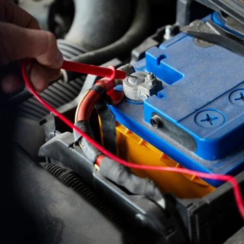 A hand is attaching a red cable to the terminal of a blue car battery in an open engine compartment, showcasing expert auto repair in Surrey and Delta.