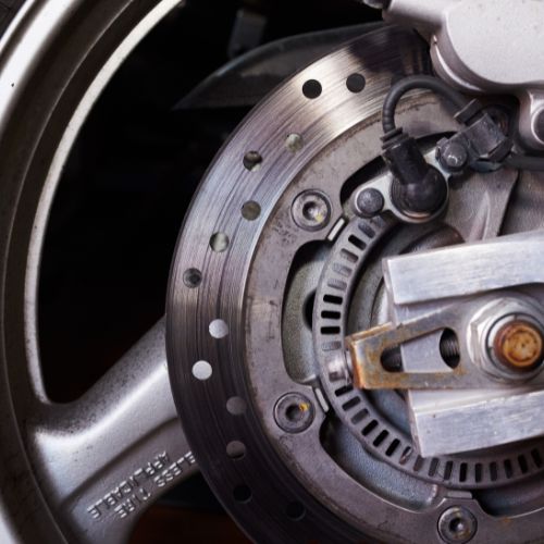 Close-up of a motorcycle disc brake, showing a metal rotor with drilled holes, caliper, and attached wiring on a wheel hub.