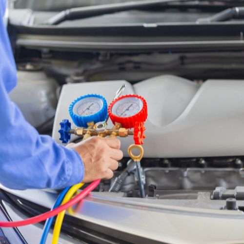 A person uses an AC manifold gauge set with red and blue hoses to check a car's air conditioning system.