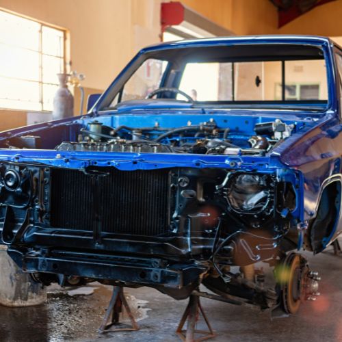 A blue car with its front end disassembled and raised on stands inside a garage.