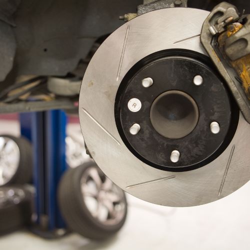 Close-up of a car brake disc on a lifted vehicle in a garage. Wheels and other equipment are visible in the background.