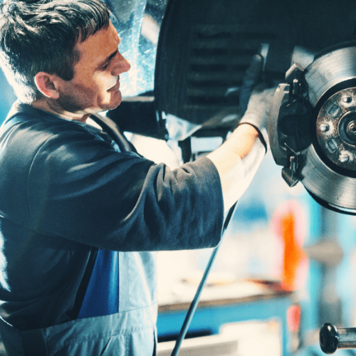 Mechanic working on a car brake system, inspecting parts while wearing gloves and a work apron.