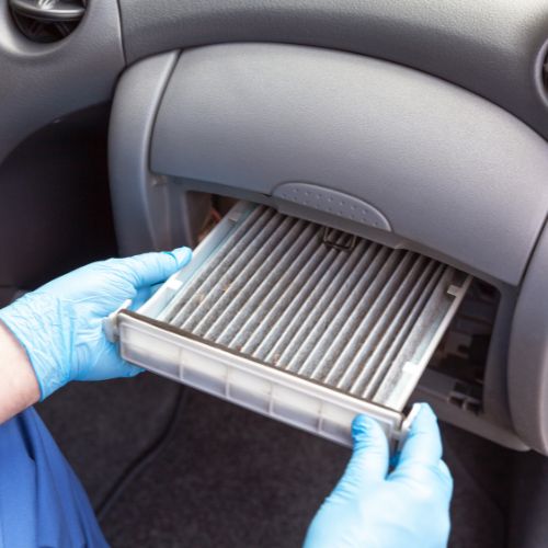 A person wearing blue gloves removes a cabin air filter from a vehicle's glove compartment.