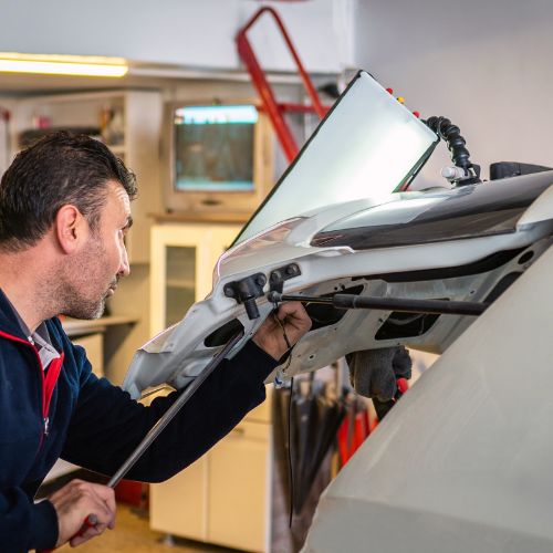 Mechanic repairing a car hood in a garage, using a tool to make adjustments.