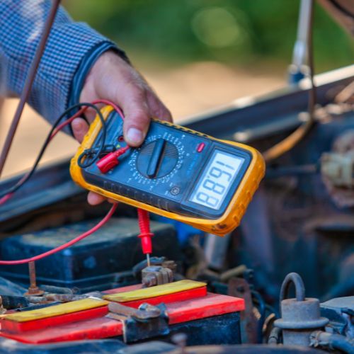 A person uses a digital multimeter to test a car battery's voltage under the hood.