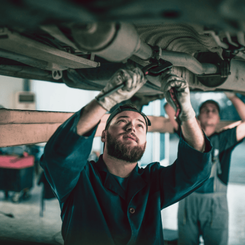 Two mechanics, one in focus, work under a vehicle. Both are wearing gloves and concentrating on repairs in a well-lit garage.