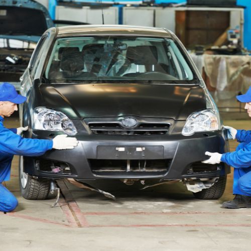 Two mechanics in blue uniforms work on the front bumper of a black car in an auto repair shop.