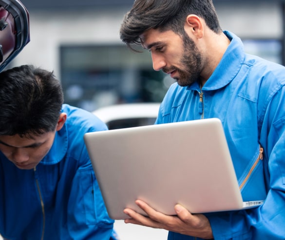 Two mechanics in blue uniforms work on a car, one holding a laptop while the other examines the engine.