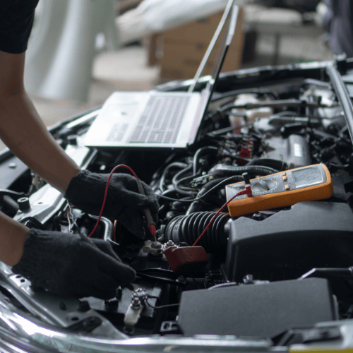 A person wearing gloves uses diagnostic tools on a car engine with an open hood. A laptop and an orange multimeter are on the engine.