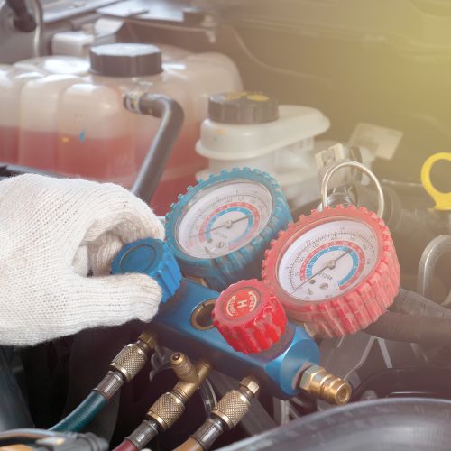 A person wearing a glove adjusts a manifold gauge set on an air conditioning system, with car engine components visible in the background.