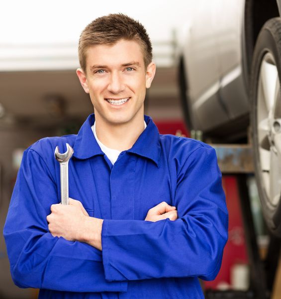 A mechanic in a blue uniform holding a wrench stands in front of a vehicle on a lift.