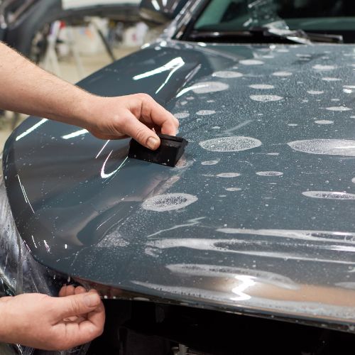 Person applying a protective film to a car hood with a squeegee tool.
